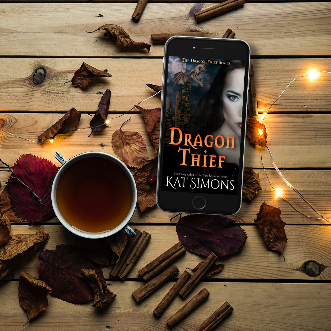 Cover of Dragon Thief book on phone laying on wood background with cup of tea, flowers and cinnamon sticks